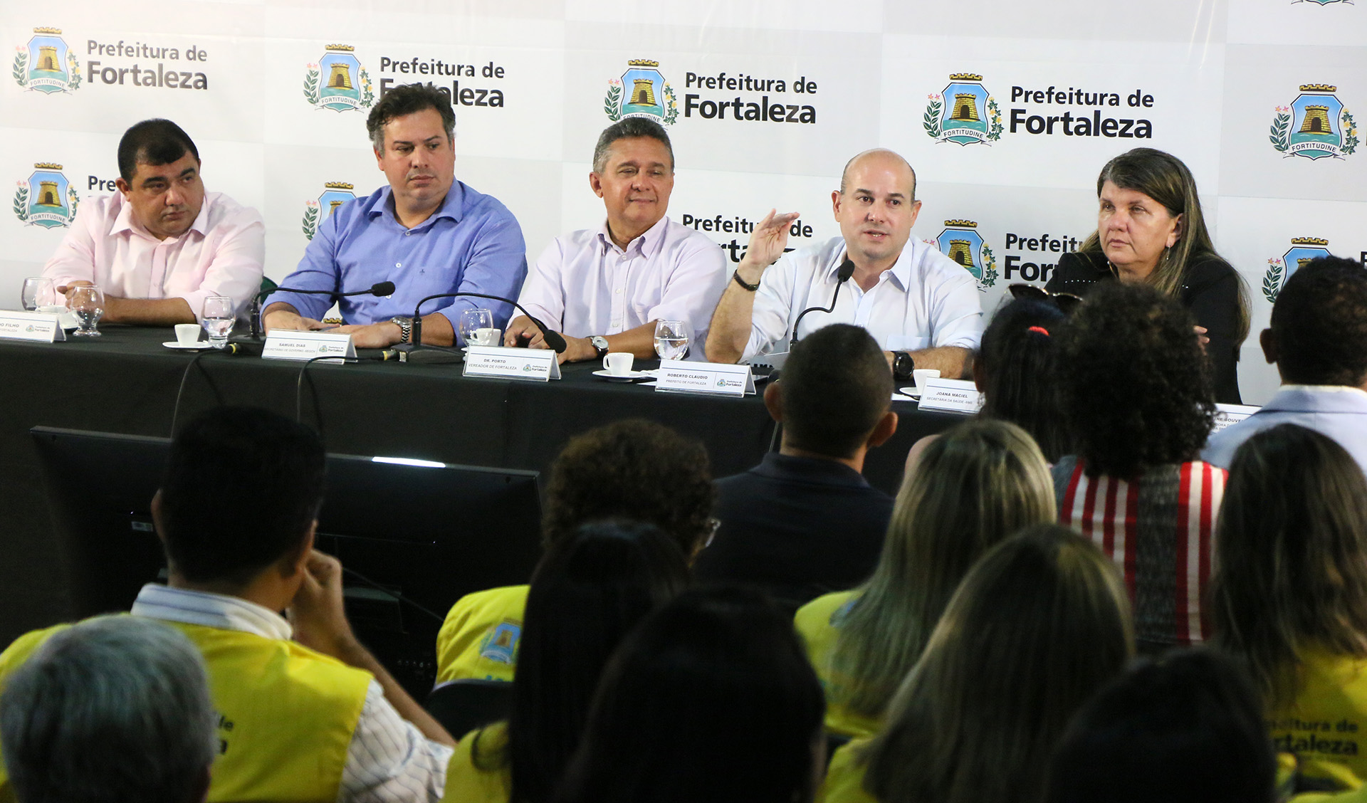 prefeito sentado à mesa falando ao microfone ao lado de secretários sentados e pessoas assistindo em frente